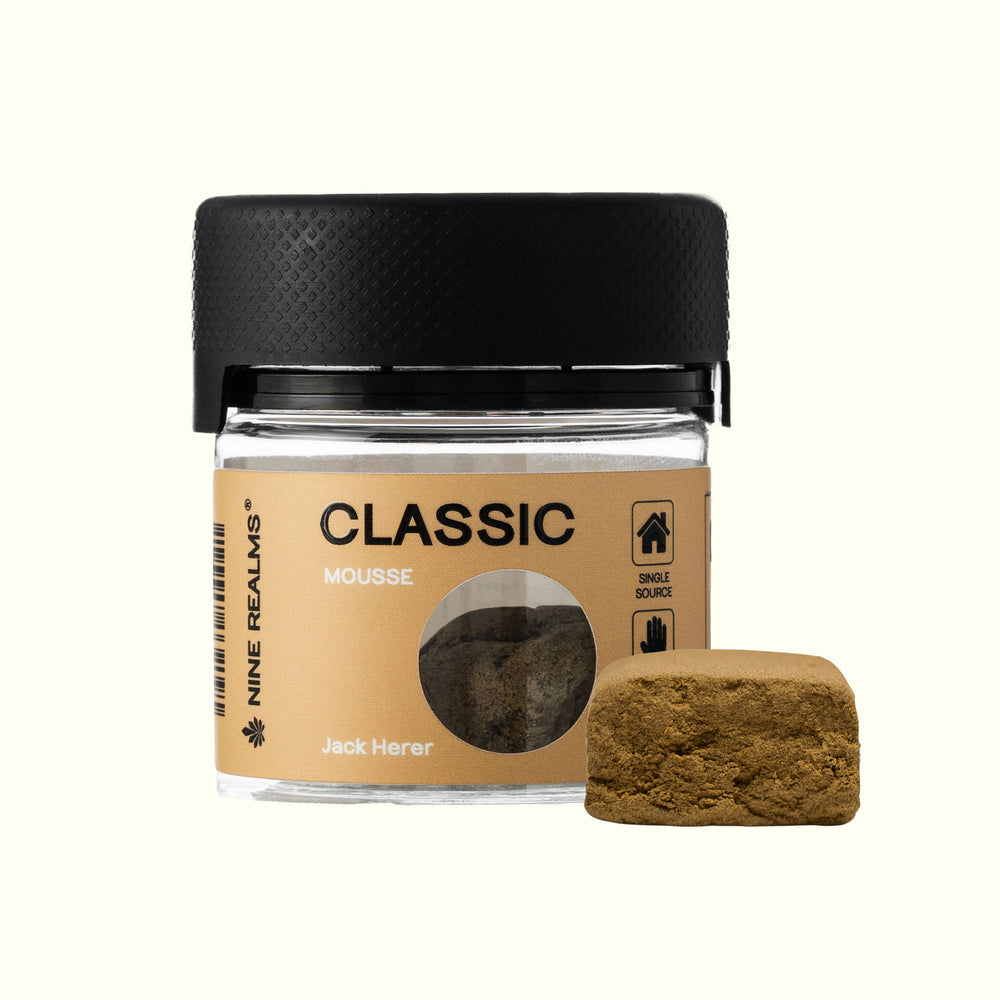 A small clear jar of Nine Realms Classic La Mousse Hash with a black lid, shown next to a textured square sample of the brownish-green bait on a light cream background.