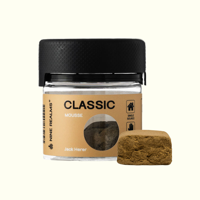 A small clear jar of Nine Realms Classic La Mousse Hash with a black lid, shown next to a textured square sample of the brownish-green bait on a light cream background.